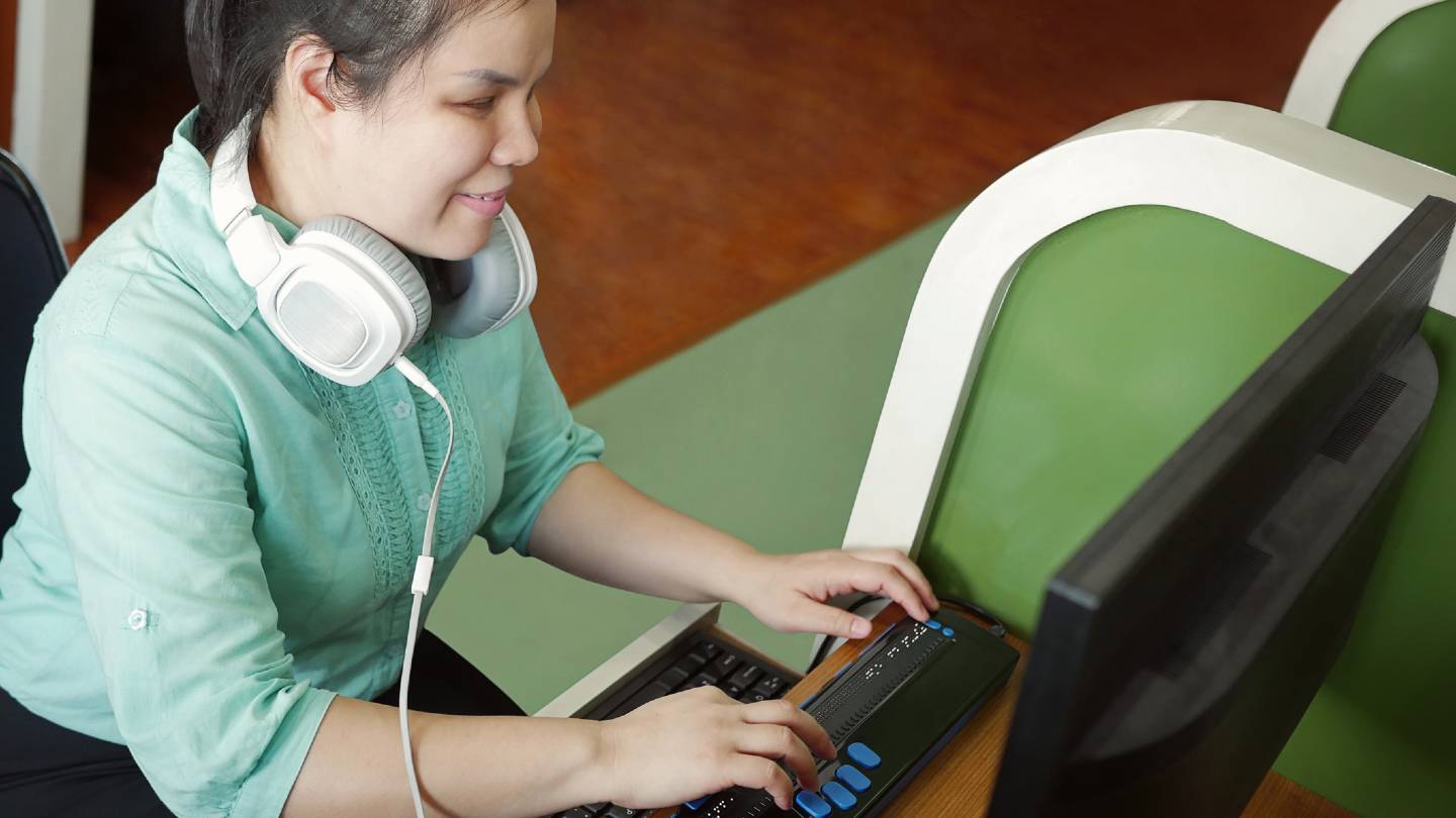 Young blind woman with headphones using computer with refreshable braille display or braille terminal, a technology device for persons with visual disabilities.