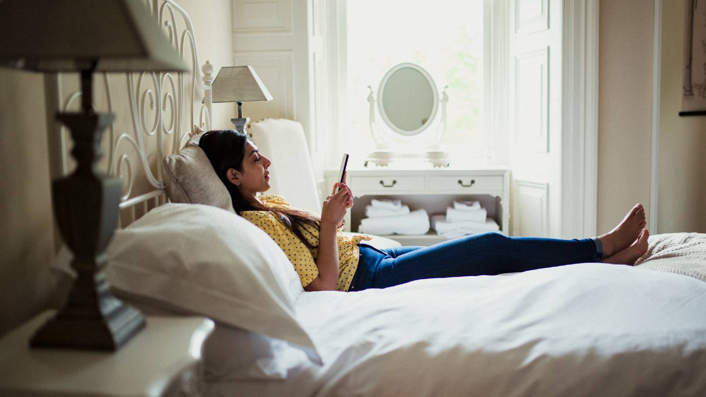 Woman relaxing on hotel bed looking at her phone.